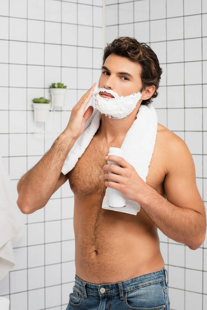 Young shirtless man with towel applying shaving foam and looking at camera in bathroom  - Foto, afbeelding