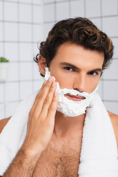 Brunette man with towel around neck applying shaving foam  - Foto, Bild