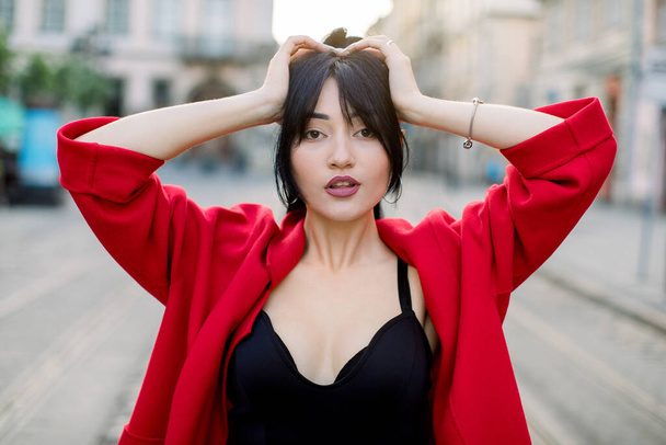 Outdoor portrait of young attractive confident Asian woman, wearing black top and stylish red blazer, posing on the street of old European city, with arms raised to her head. Urban fashion - Photo, Image