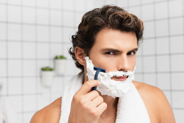 Young man with shaving foam on face holding disposable razor in bathroom  - Фото, изображение