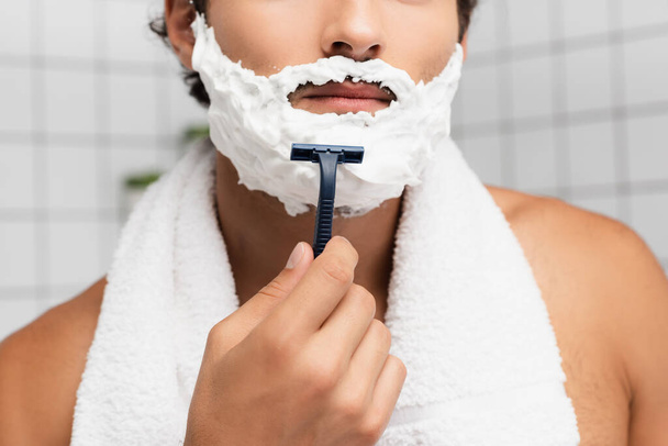 Cropped view of young man with shaving foam on face and towel around neck holding razor  - Foto, Imagem