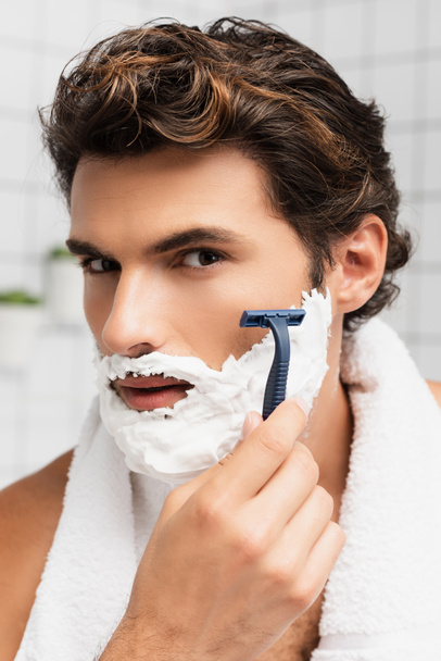 Brunette man with shaving foam on face shaving with razor in bathroom  - Φωτογραφία, εικόνα