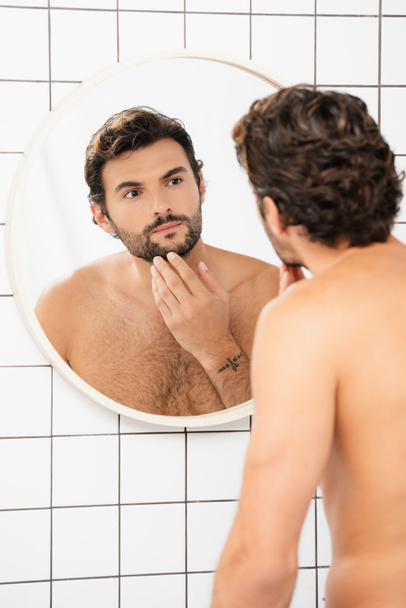 hombre sin camisa tocando la barbilla mientras mira el espejo en el baño  - Foto, imagen