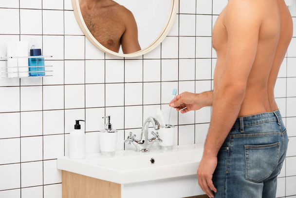 Обрезанный вид человека без рубашки, берущего зубную щетку из туалетных принадлежностей в ванной комнате  - Фото, изображение