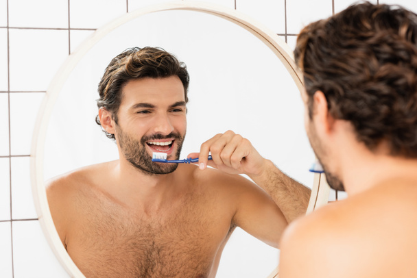 Lächelnder Mann ohne Hemd reflektiert im Spiegel beim Zähneputzen im Badezimmer  - Foto, Bild