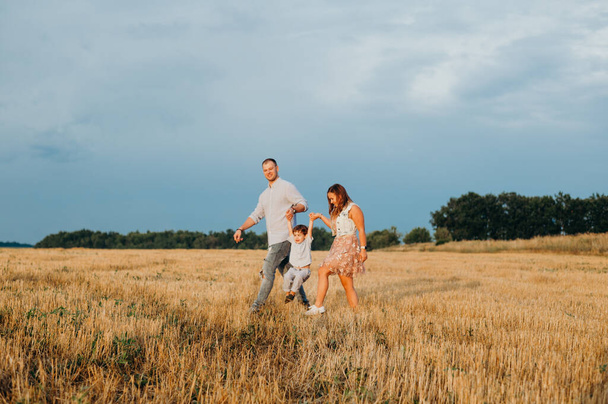 The concept of a happy family. Son with mom and daddy. Happy family in the field evening light of a sun. Mother, father and baby son happy walk at sunset, parents hold the baby.  - 写真・画像