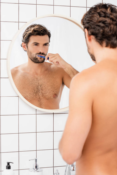 Barded i bez koszuli człowiek patrząc w lustro podczas mycia zębów na rozmytym pierwszym planie w łazience  - Zdjęcie, obraz