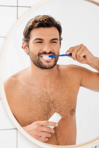 Muscular man holding tube with toothpaste while brushing teeth near mirror  - Fotó, kép