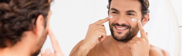 Hombre sonriente aplicando crema cosmética en la cara cerca del espejo en primer plano borroso, pancarta  - Foto, imagen