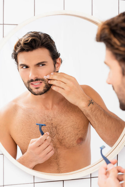 Shirtless man touching cheek with wound and holding razor near mirror on blurred foreground  - Photo, image