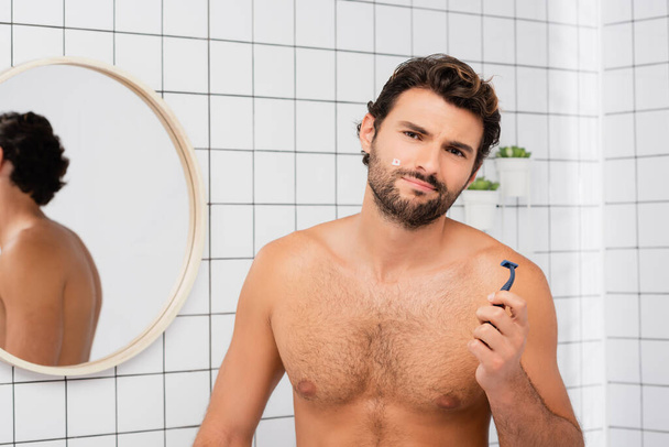 Hombre sin camisa con herida en la mejilla mirando a la cámara mientras sostiene la navaja en el baño  - Foto, imagen