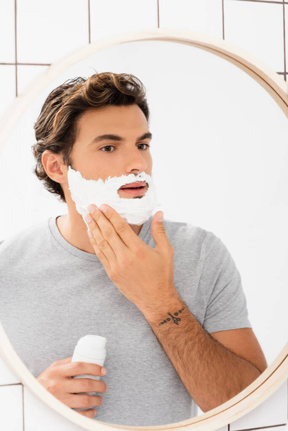 Young man applying shaving foam near mirror in bathroom  - Zdjęcie, obraz