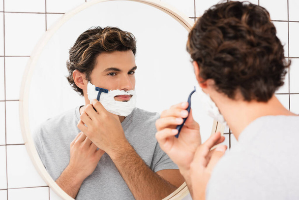 Young man looking at mirror while shaving on blurred foreground in bathroom  - Valokuva, kuva