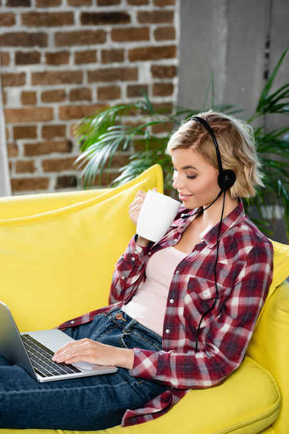 young blonde woman having webinar sitting at home and drinking tea - Photo, Image