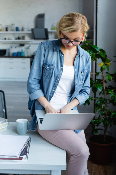 young blonde woman in eyeglasses working from home sitting on table - Photo, Image
