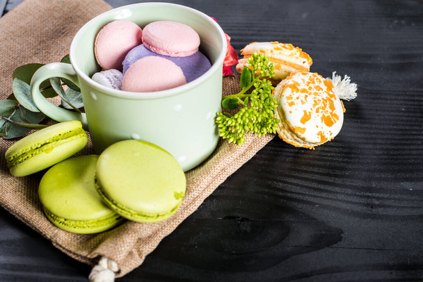 Sweet colored macaroons in composition with flowers on black background. Copy space - Фото, зображення