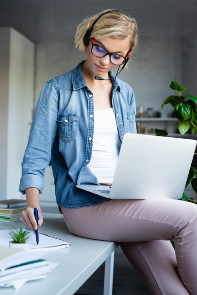 young blonde woman in eyeglasses working from home with laptop and writing notes - Foto, Imagen