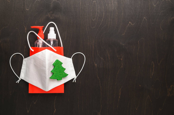 Bolsa roja con botellas de desinfectante y una máscara protectora reutilizable, decorada con árbol de Navidad sobre un fondo marrón oscuro. - Foto, imagen