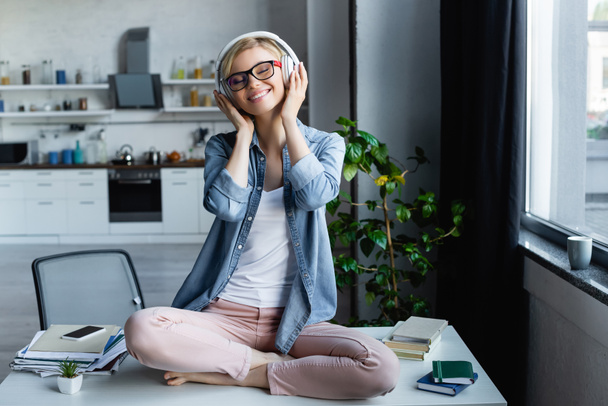 young blonde woman in eyeglasses listening music and sitting on table - Фото, изображение