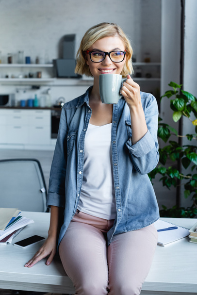 Lächelnde blonde Frau mit Brille, die zu Hause Tee trinkt - Foto, Bild