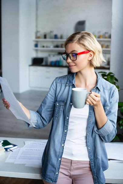young blonde woman in eyeglasses looking at document and drinking tea - Fotó, kép