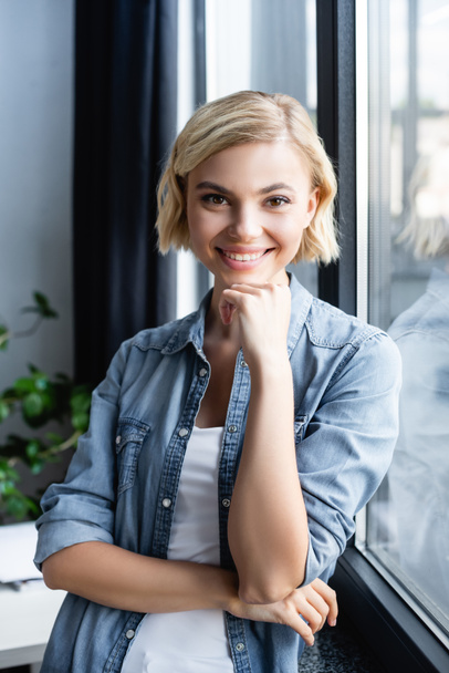 portrait of smiling blonde woman standing near window - Photo, Image
