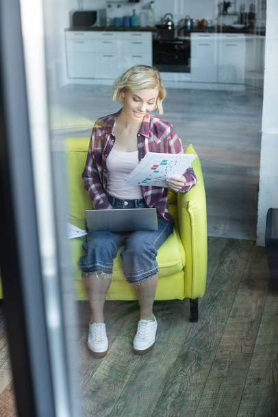vue à travers le verre de femme blonde en chemise à carreaux travaillant à la maison et en regardant à travers les documents - Photo, image