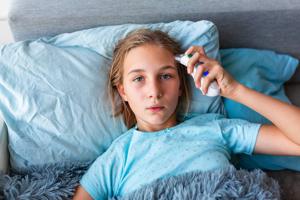 Sick teenage little girl with high fever and headache laying in bed checking her temperature. Stay at home during corona virus epidemic if you feel sick - Φωτογραφία, εικόνα