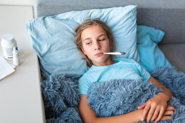 Sick teenage little girl with high fever and headache laying in bed and holding thermometer in her mouth. Stay at home during corona virus epidemic if you feel sick - Foto, Imagen