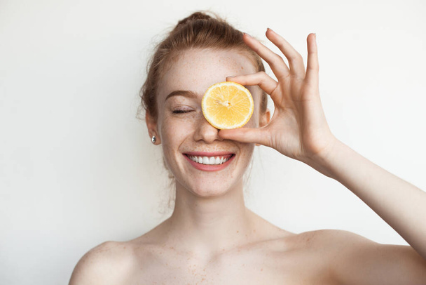 Red haired woman with freckles covering an eye with a lemon and smile with naked shoulders on a white studio wall - Foto, immagini