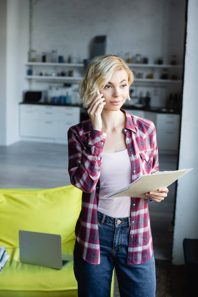 young blonde woman in checkered shirt holding documents and talking on smartphone - Foto, afbeelding