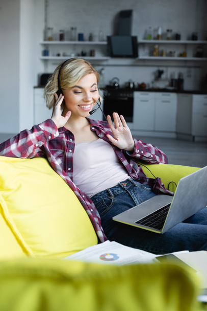 junge blonde Frau mit Kopfhörer bei einer Online-Konferenz und winkender Hand - Foto, Bild