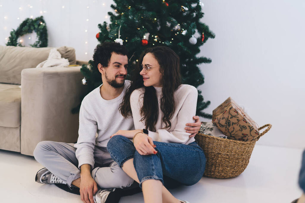 Positives junges Paar sitzt neben geschmücktem Weihnachtsbaum zu Hause und schaut sich an, kaukasische Liebhaber in Freizeitkleidung genießen Winterurlaub und Neujahrsfeier im gemütlichen Heim-Interieur - Foto, Bild