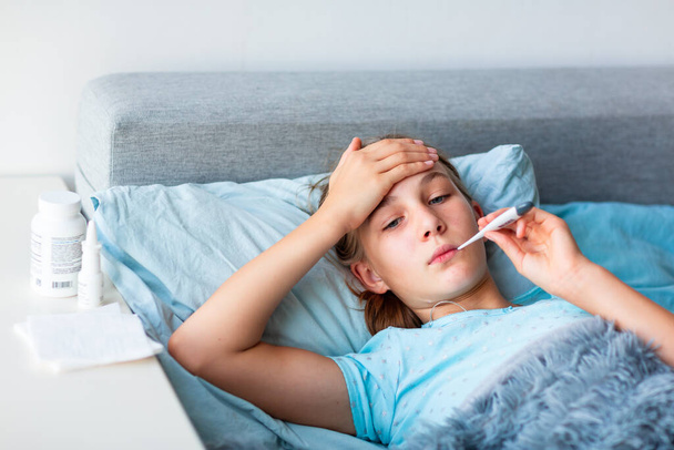 Sick teenage little girl with high fever and headache laying in bed and holding thermometer in her mouth. Stay at home during corona virus epidemic if you feel sick - Φωτογραφία, εικόνα