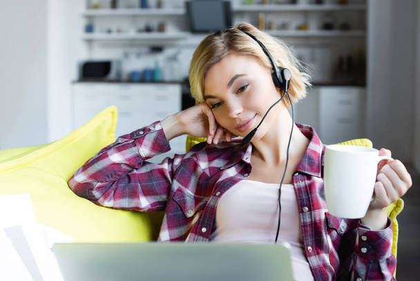 young blonde woman in headphones working from home and drinking tea - Fotó, kép
