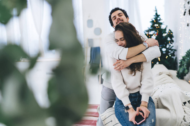 pozytywny kaukaski mężczyzna przytulając jego dziewczyna czule korzystających Boże Narodzenie święto w świątecznym urządzone mieszkanie, romantyczna para w miłość uczucie szczęścia razem w przytulne wnętrze domu - Zdjęcie, obraz
