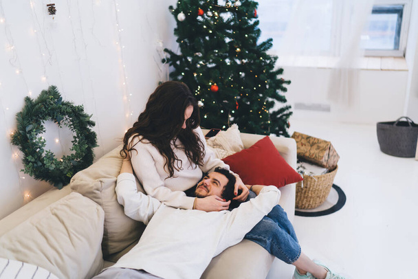 Young woman in casual clothing sitting on sofa with boyfriend lying on laps in cosy room decorated in neutral with Christmas tree and lights - Zdjęcie, obraz