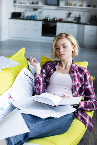 молодая блондинка в клетчатой рубашке лежит на диване с документами и держит блокнот - Фото, изображение