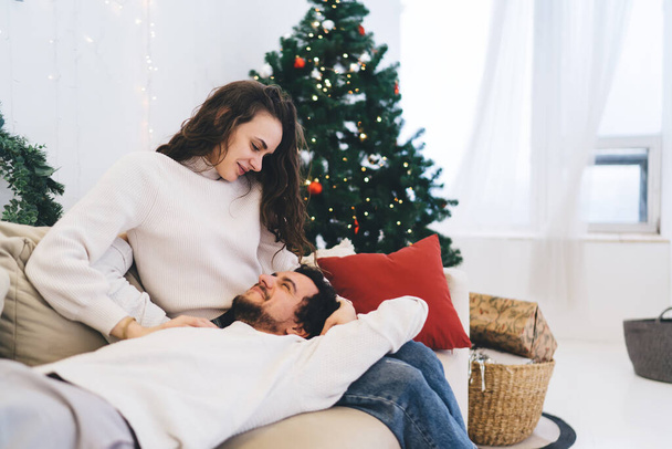 Gündelik giysiler içindeki genç adam kız arkadaşının kucağına uzanmış ve gülümserken evde Noel boyunca sevgi ve şefkatle gözlerinin içine bakıyordu. - Fotoğraf, Görsel