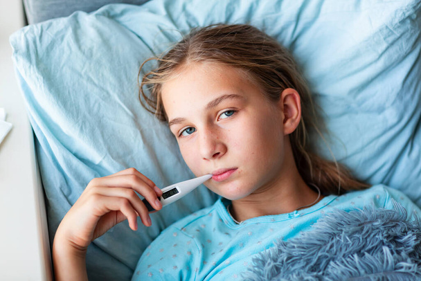 Ziek tienermeisje met hoge koorts en hoofdpijn in bed en thermometer in haar mond. Blijf thuis tijdens de corona-virusepidemie als u zich ziek voelt - Foto, afbeelding