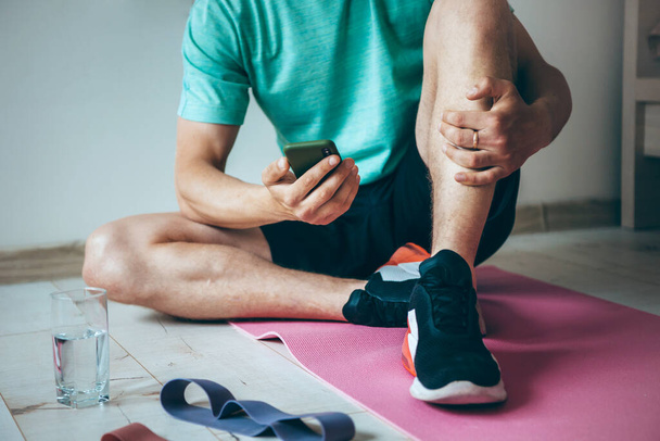 Homem caucasiano sentado no chão enquanto relaxa depois de fazer fitness em casa está usando um telefone e um copo de água - Foto, Imagem