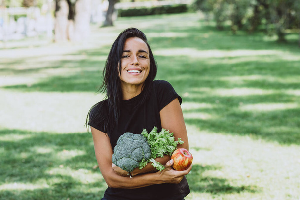 Porträt einer lächelnden jungen, braun gebrannten Frau im Park, die einen Apfel und einen Strauß Grünzeug mit Brokkoli in der Hand hält, Ernährung, gesunde Ernährung. Weiche selektive Fokussierung. - Foto, Bild