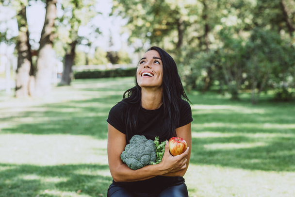 Porträt einer lächelnden jungen, braun gebrannten Frau im Park, die einen Apfel und einen Strauß Grünzeug mit Brokkoli in der Hand hält, Ernährung, gesunde Ernährung. Weiche selektive Fokussierung. - Foto, Bild