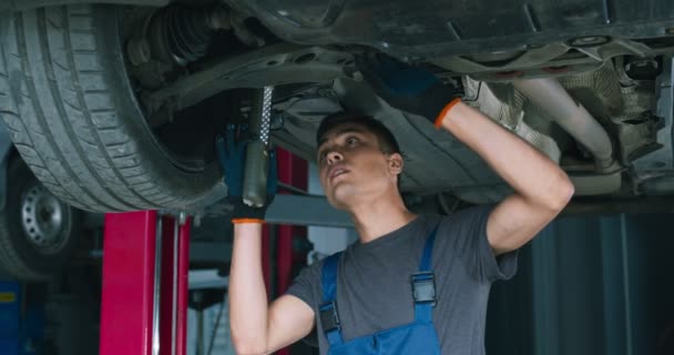 Νεαρός μηχανικός επισκευή ανάρτησης αυτοκινήτου, στέκεται κάτω από το όχημα στο γκαράζ - Πλάνα, βίντεο