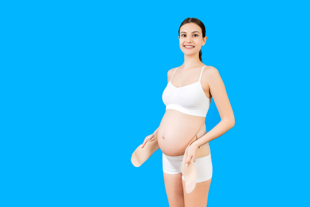 Portrait of pregnant woman in underwear dressing maternity belt against pain in the back at blue background with copy space. Orthopedic abdominal support belt concept. - Photo, Image