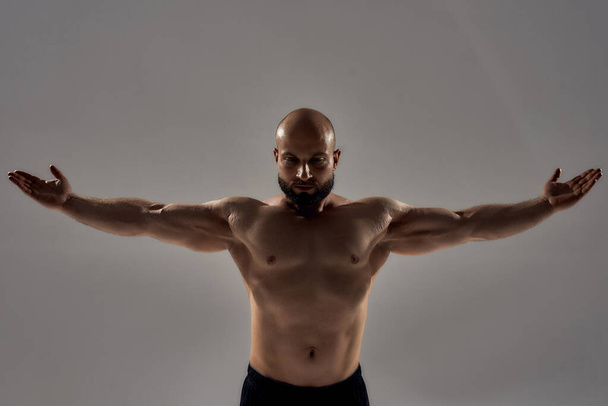 Professional bodybuilder posing. Muscular strong bald man with naked torso standing with outstretched arms isolated over grey background - Фото, изображение