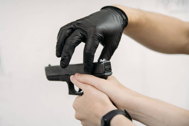 Vivir en una pandemia. Instructor de tiro en guantes que muestra al cliente cómo usar un arma en un campo de tiro en medio de una pandemia - Foto, imagen