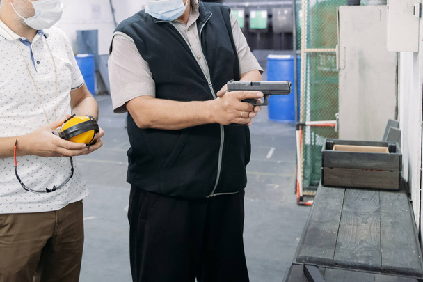Leven in een pandemie. Schietinstructeur in een medisch masker en handschoenen die de cliënt laat zien hoe hij een pistool moet gebruiken op een schietbaan te midden van een pandemie - Foto, afbeelding