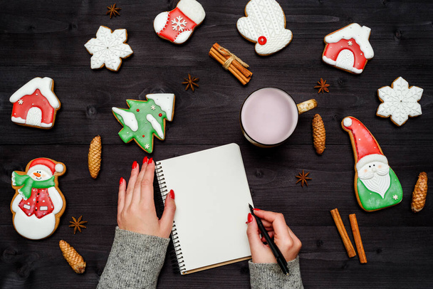 Woman with red manicure writing New year resolutions in blank notebook, copy space. Christmas background with gingerbread cookies, cinnamon and pine cones on dark wooden desk. Top view, flat lay - Zdjęcie, obraz