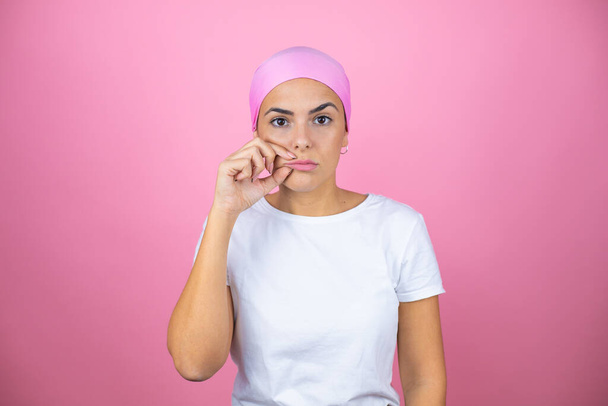 Fiatal gyönyörű nő visel rózsaszín fejkendő felett elszigetelt rózsaszín háttér száj és ajkak zárva, mint cipzár ujjakkal. Titkos és csendes, tabu beszéd - Fotó, kép